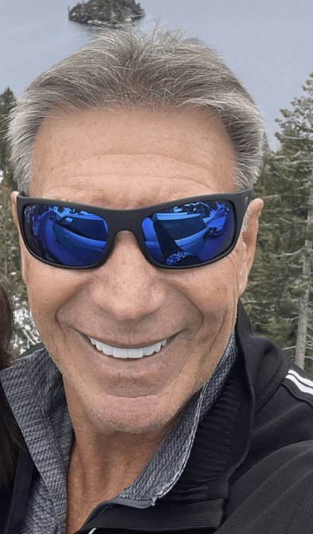 Joyful man outdoors in nature, wearing sunglasses, smiling by serene water and trees.